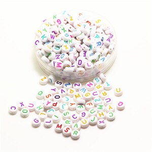 May include: A clear plastic container overflowing with white, round beads, each displaying a letter of the alphabet in various colours. Some beads are scattered on a white surface. The letters are in a variety of colours.