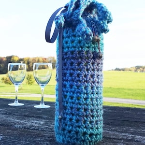 May include: A crocheted wine bottle cozy in shades of blue and purple with a gray ribbon and beaded embellishments. The cozy is sitting on a wooden table with two wine glasses in the background.