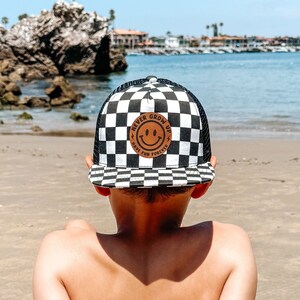 May include: A young person wearing a black and white checkered baseball cap with a brown patch that says "Never Grow Up" and a smiley face. The person is sitting on a sandy beach with their back to the camera, looking out at the ocean.