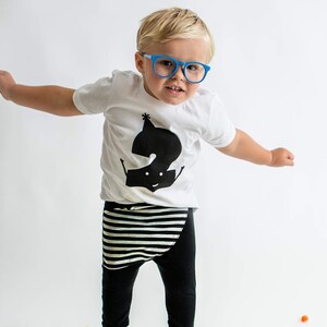 Peut inclure: Un tout-petit portant un t-shirt blanc avec un graphique noir repr&eacute;sentant le chiffre "2" et une ceinture noire et blanche ray&eacute;e sur un pantalon noir. Le tout-petit porte des lunettes bleues et des chaussures bleues et saute en l'air. Il y a des pompons color&eacute;s &eacute;parpill&eacute;s sur le sol.