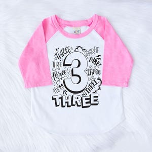 May include: A white and pink baseball tee shirt with a black graphic that says "Three" with the number 3 in the center. The number 3 is surrounded by the word "Three" written multiple times.