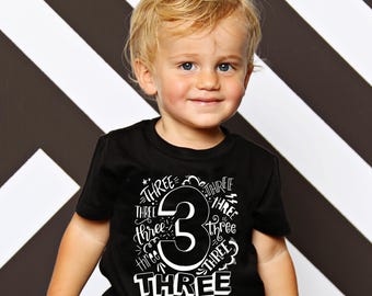 May include: A young child wearing a black t-shirt with a large white number "3" and the word "THREE" repeated around it. The child is also wearing black and white striped pants. The background is a black and white chevron pattern.