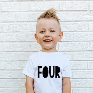 May include: A young boy wearing a white t-shirt with the word "FOUR" printed in black. He is wearing blue denim shorts and black sneakers. He is standing in front of a white brick wall.
