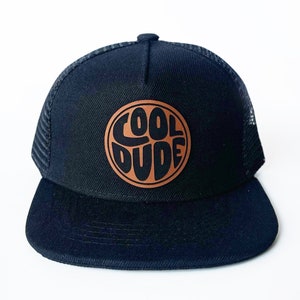 May include: Black trucker hat with a curved brim and mesh back. The hat features a circular brown patch with the words "COOL DUDE" in a bold, rounded font. The hat is set against a white background.