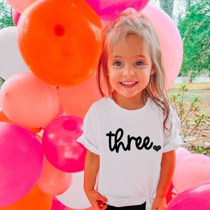 Puede incluir: Una ni&ntilde;a lleva una camiseta blanca con la palabra "three" en escritura negra y un peque&ntilde;o coraz&oacute;n negro. La ni&ntilde;a lleva leggings negros y zapatillas blancas, de pie frente a una colorida exhibici&oacute;n de globos rosas, naranjas y blancos.