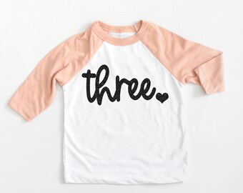 May include: White long-sleeve shirt with pink sleeves and black text that says "three" with a small black heart.