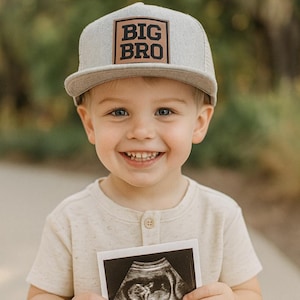 May include: A smiling child wearing a gray cap with "BIG BRO" printed on it, holding an ultrasound image. The child is wearing a light-colored shirt. The background is blurred with green foliage.