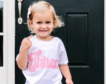 May include: A young child wearing a white t-shirt with the word "Three" in pink script. The child is wearing denim shorts and is standing on a brick step.