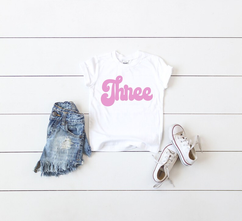 May include: White t-shirt with pink "Three" text in a retro font. The shirt is paired with denim shorts and white sneakers.