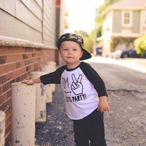 May include: A young child wearing a black and white baseball tee shirt with the text "I'm  --- Let's Party!" and a peace sign graphic. The child is wearing a camouflage baseball cap and black pants.