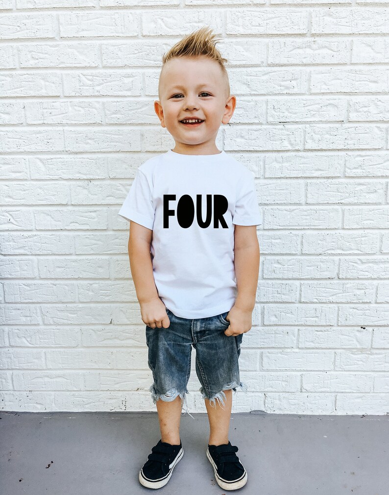 May include: A young boy wearing a white t-shirt with the word "FOUR" printed in black. He is wearing blue denim shorts and black sneakers. He is standing in front of a white brick wall.