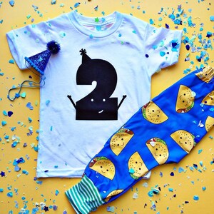 Peut inclure: Un t-shirt blanc avec un num&eacute;ro deux noir dessus. Le num&eacute;ro deux porte un chapeau de f&ecirc;te et a un visage souriant. Le t-shirt est associ&eacute; &agrave; un pantalon bleu avec un motif de tacos jaunes. Le pantalon a un ourlet ray&eacute; vert et blanc.