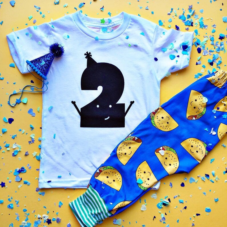 Peut inclure: Un t-shirt blanc avec un num&eacute;ro deux noir dessus. Le num&eacute;ro deux porte un chapeau de f&ecirc;te et a un visage souriant. Le t-shirt est associ&eacute; &agrave; un pantalon bleu avec un motif de tacos jaunes. Le pantalon a un ourlet ray&eacute; vert et blanc.