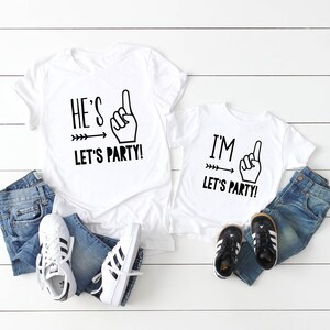 May include: Two white t-shirts with black text and graphics. The larger shirt says "He's 1 Let's Party!" and the smaller shirt says "I'm 1 Let's Party!"