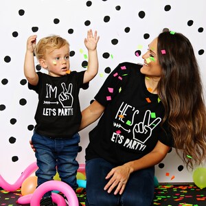 May include: A mother and child wearing matching black t-shirts with white text that says "I'm Two Let's Party!" and "He's Two Let's Party!" They are standing in front of a white background with black polka dots. Confetti is falling around them.