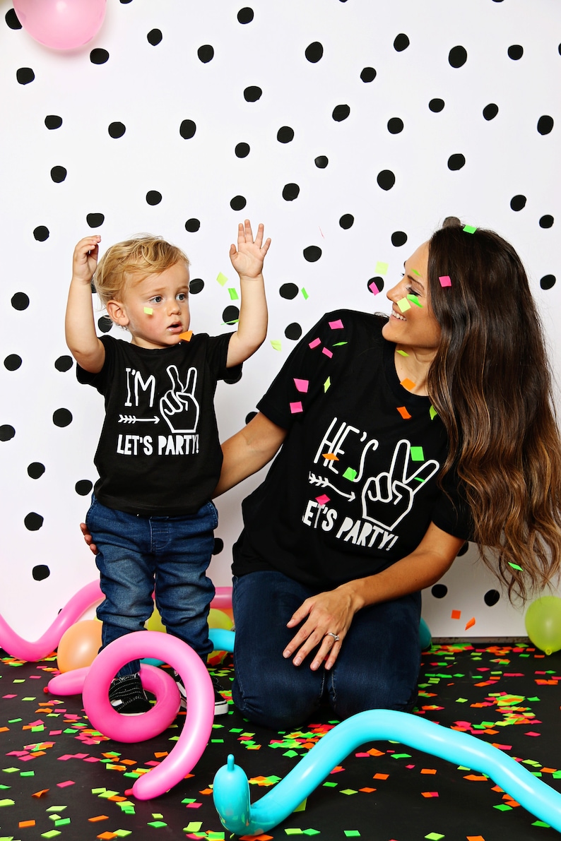 May include: A mother and child wearing matching black t-shirts with white text that says "I'm Two Let's Party!" and "He's Two Let's Party!" They are standing in front of a white background with black polka dots. Confetti is falling around them.