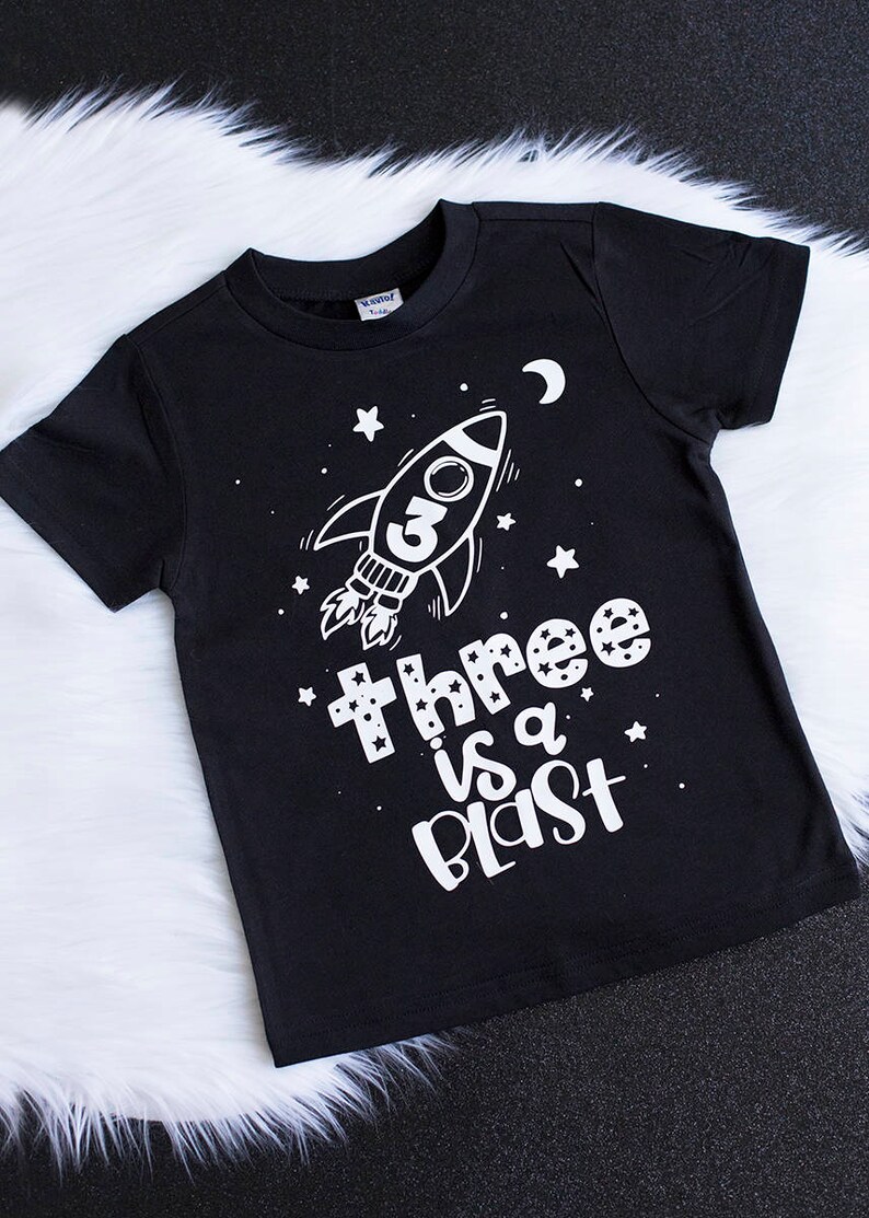May include: Black t-shirt with a white rocket ship and stars. The text "Three is a Blast" is printed on the shirt.