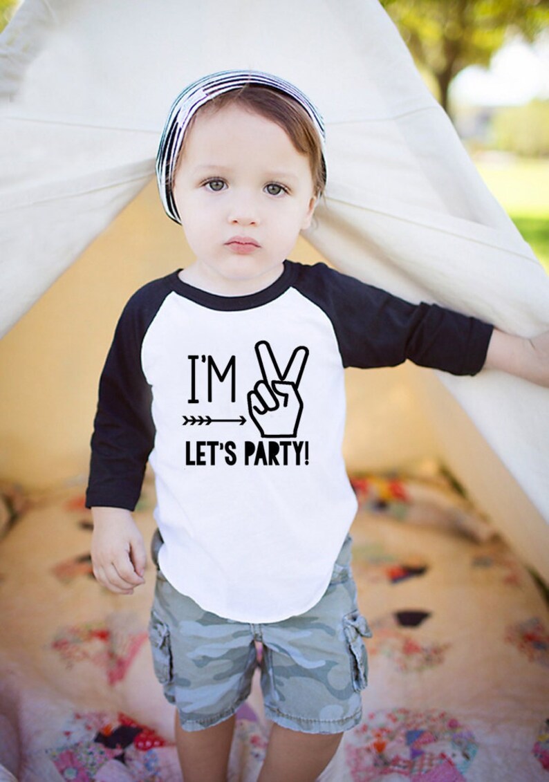 May include: A toddler wearing a white and black baseball tee shirt with the text "I'm Two Let's Party!" and a peace sign graphic. The toddler is wearing camouflage shorts and a striped hat.