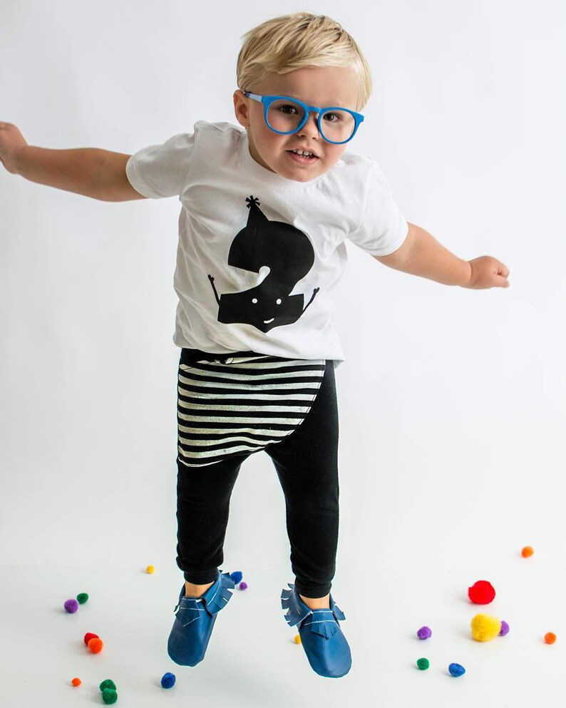Peut inclure: Un tout-petit portant un t-shirt blanc avec un graphique noir repr&eacute;sentant le chiffre "2" et une ceinture noire et blanche ray&eacute;e sur un pantalon noir. Le tout-petit porte des lunettes bleues et des chaussures bleues et saute en l'air. Il y a des pompons color&eacute;s &eacute;parpill&eacute;s sur le sol.