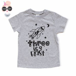 May include: Gray short-sleeved t-shirt with a black rocket ship graphic and the text "Three is a Blast".