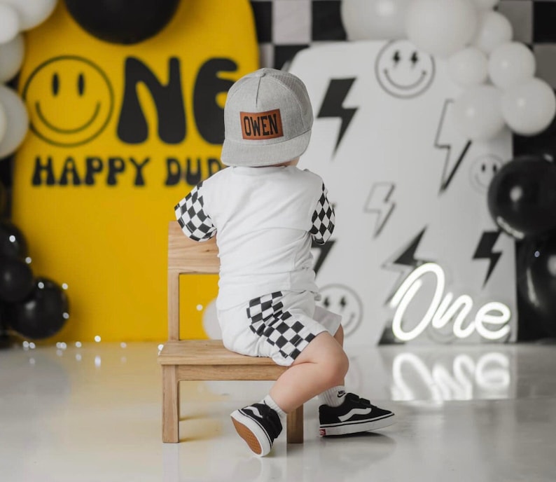 May include: A young child wearing a black and white checked shirt and black and white checked shorts sits on a wooden stool. The child is wearing a black and white baseball cap with the name "Owen" on it. The child is sitting in front of a yellow and black backdrop with the words "One Happy Dude" on it.