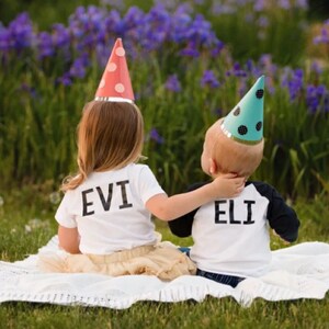 May include: Two children wearing party hats are sitting on a white blanket. The girl is wearing a white shirt with the name "EVI" on the back. The boy is wearing a white shirt with the name "ELI" on the back.