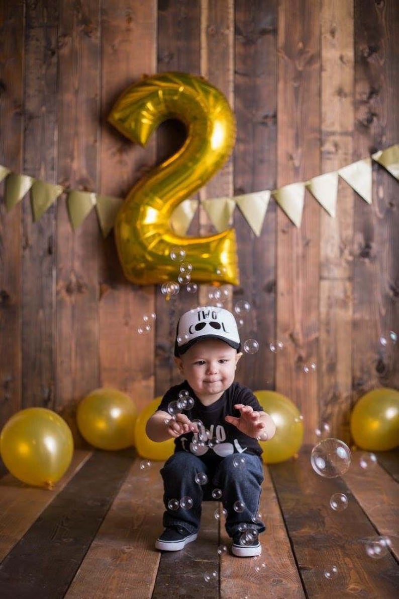 May include: A toddler wearing a black shirt with the text "Two Cool" and a white trucker hat with the text "Two Cool" sits in front of a gold number two balloon. The toddler is surrounded by gold balloons and bubbles.