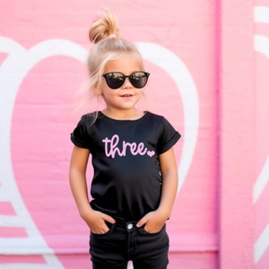 Puede incluir: Una ni&ntilde;a peque&ntilde;a con una camiseta negra con la palabra "three" en letras rosas y un coraz&oacute;n rosa. Lleva gafas de sol negras y pantalones vaqueros negros. La ni&ntilde;a est&aacute; de pie frente a una pared rosa con corazones blancos.