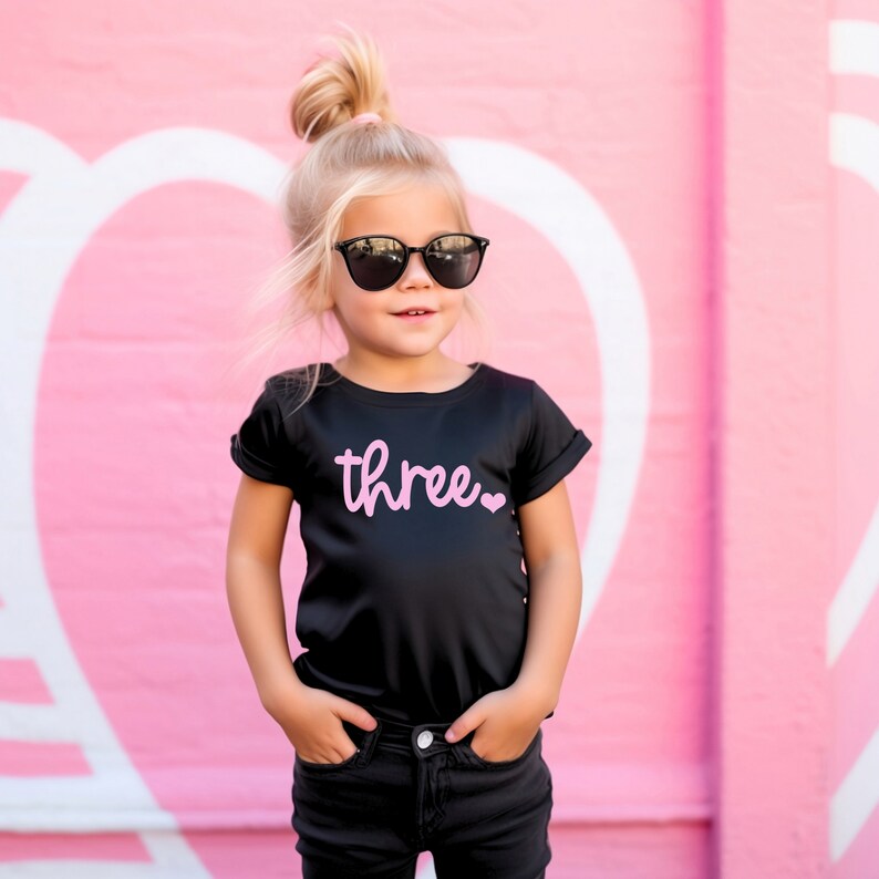 Puede incluir: Una ni&ntilde;a peque&ntilde;a con una camiseta negra con la palabra "three" en letras rosas y un coraz&oacute;n rosa. Lleva gafas de sol negras y pantalones vaqueros negros. La ni&ntilde;a est&aacute; de pie frente a una pared rosa con corazones blancos.