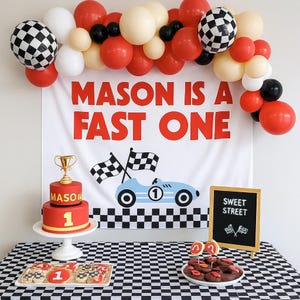 May include: A racing-themed party setup with a banner reading "MASON IS A FAST ONE." Includes a red cake with a gold trophy, cookies, and a black and white checkered tablecloth. Balloons in red, white, black, and cream colors complete the scene.
