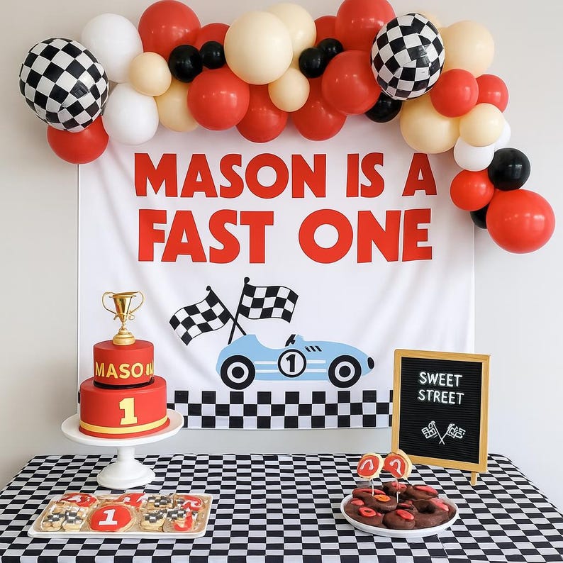 May include: A racing-themed party setup with a banner reading "MASON IS A FAST ONE." Includes a red cake with a gold trophy, cookies, and a black and white checkered tablecloth. Balloons in red, white, black, and cream colors complete the scene.