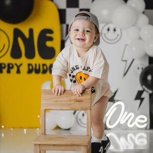 May include: A young child wearing a grey baseball cap and a white t-shirt with a yellow smiley face graphic and the text "One Lap" is standing next to a wooden chair. The child is smiling and looking at the camera. The background is decorated with black and white chequered patterns, yellow and white balloons, and a neon sign that says "One".