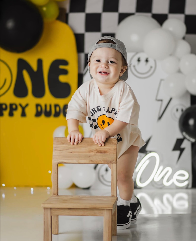 May include: A young child wearing a grey baseball cap and a white t-shirt with a yellow smiley face graphic and the text "One Lap" is standing next to a wooden chair. The child is smiling and looking at the camera. The background is decorated with black and white chequered patterns, yellow and white balloons, and a neon sign that says "One".