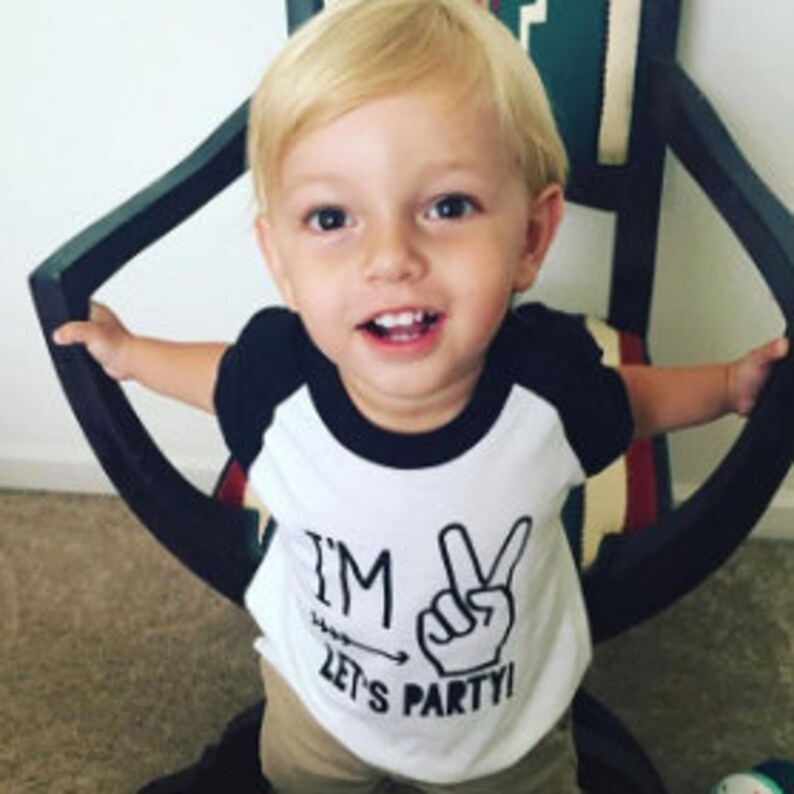 May include: A white and black toddler t-shirt with a black and white graphic that says "I'm 2 Let's Party!" with a peace sign and an arrow pointing to the peace sign.