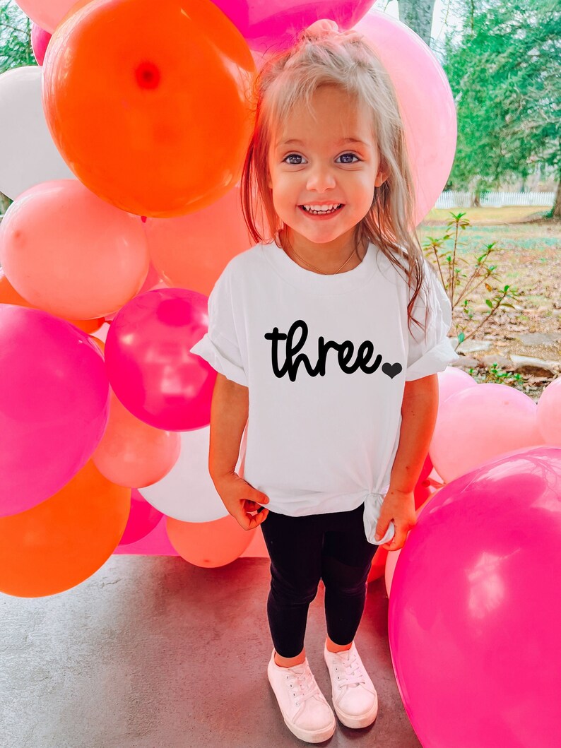 Puede incluir: Una ni&ntilde;a lleva una camiseta blanca con la palabra "three" en escritura negra y un peque&ntilde;o coraz&oacute;n negro. La ni&ntilde;a lleva leggings negros y zapatillas blancas, de pie frente a una colorida exhibici&oacute;n de globos rosas, naranjas y blancos.