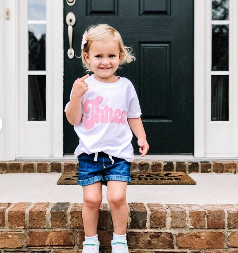 May include: A young child wearing a white t-shirt with the word "Three" in pink script. The child is wearing denim shorts and is standing on a brick step.
