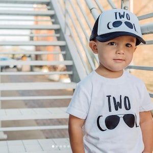 May include: A toddler wearing a white t-shirt with the text "TWO COOL" and sunglasses graphic. The toddler is also wearing a black and white trucker hat with the text "TWO COOL" and sunglasses graphic.