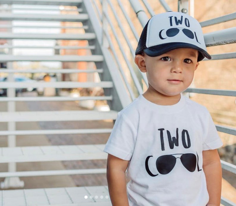 May include: A toddler wearing a white t-shirt with the text "TWO COOL" and sunglasses graphic. The toddler is also wearing a black and white trucker hat with the text "TWO COOL" and sunglasses graphic.