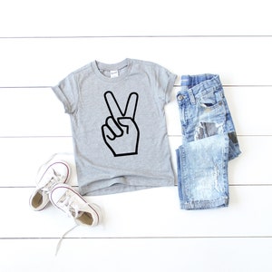 May include: A grey short-sleeved t-shirt with a black peace sign graphic. The shirt is laid flat on a white wooden surface with a pair of white trainers and a pair of blue jeans.