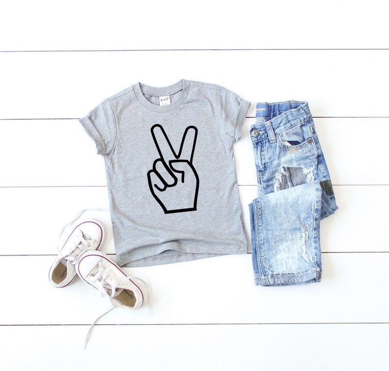 May include: A grey short-sleeved t-shirt with a black peace sign graphic. The shirt is laid flat on a white wooden surface with a pair of white trainers and a pair of blue jeans.