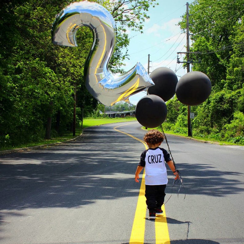 May include: A young child wearing a white t-shirt with the word "CRUZ" printed on the back walks down a paved road holding a silver number two balloon and several black balloons.