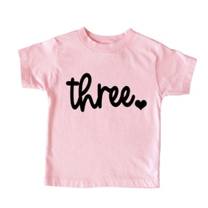 Puede incluir: Camiseta rosa claro para ni&ntilde;o con la palabra "three" en escritura negra, seguida de un peque&ntilde;o coraz&oacute;n negro. La camiseta tiene mangas cortas y cuello redondo, ideal para un tercer cumplea&ntilde;os.