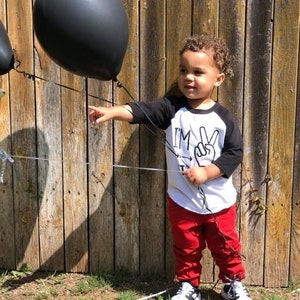 May include: A young child wearing a white and black baseball shirt with the text "I'm Two" and a peace sign. The child is holding black balloons and wearing red pants and black and white sneakers.
