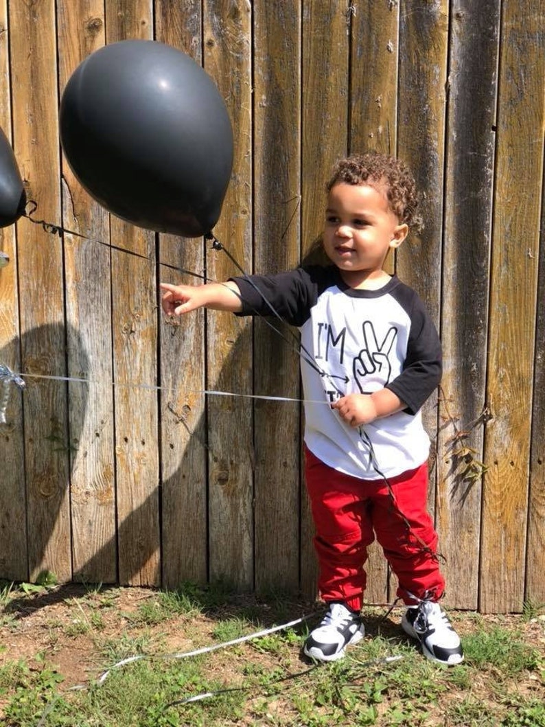 May include: A young child wearing a white and black baseball shirt with the text "I'm Two" and a peace sign. The child is holding black balloons and wearing red pants and black and white sneakers.