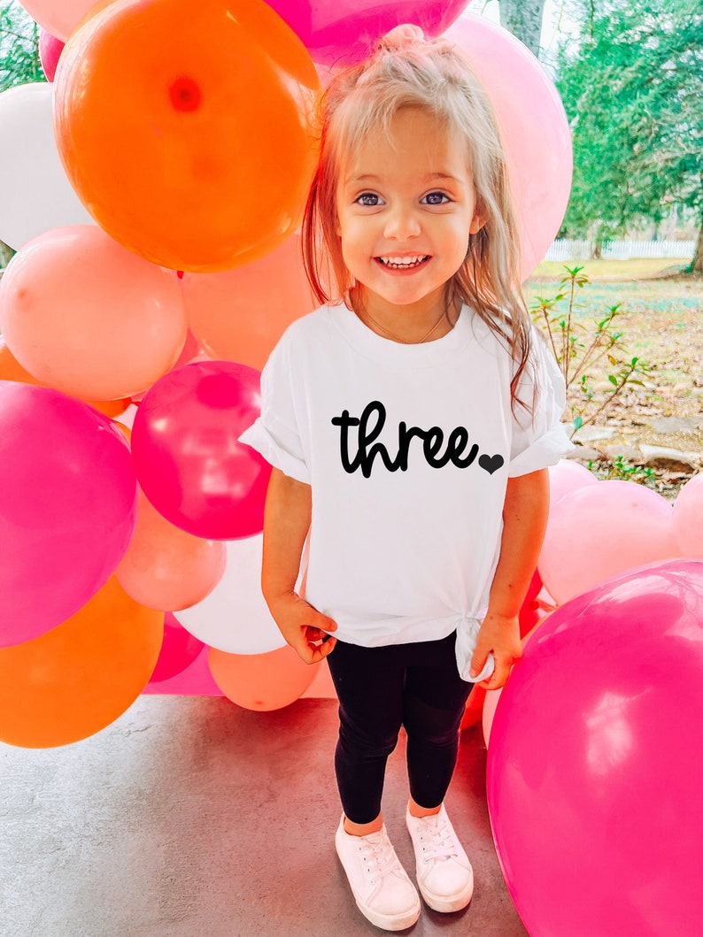 Puede incluir: Una ni&ntilde;a peque&ntilde;a lleva una camiseta blanca con la palabra "three" en escritura negra y un peque&ntilde;o coraz&oacute;n. La ni&ntilde;a est&aacute; de pie frente a una colorida exhibici&oacute;n de globos con globos rosas, naranjas y blancos. La ni&ntilde;a lleva leggings negros y zapatillas blancas.