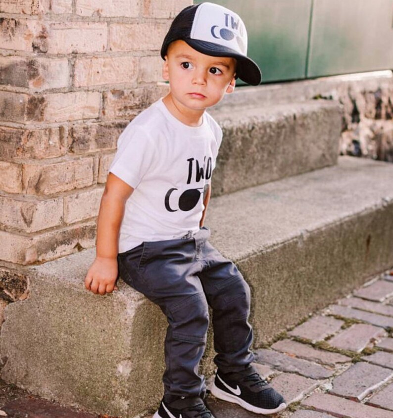 May include: A young child wearing a white t-shirt with the text "Two Cool" and black sunglasses graphic, black and white trucker hat with the text "Two Cool", black pants, and black and white sneakers.