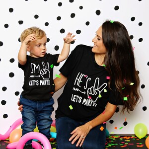 May include: A mother and child wearing matching black t-shirts with white text that says "I'm 2 Let's Party!" and "He's 2 Let's Party!" The child is holding up two fingers in a peace sign. They are standing in front of a black and white polka dot background with colorful confetti on the floor.