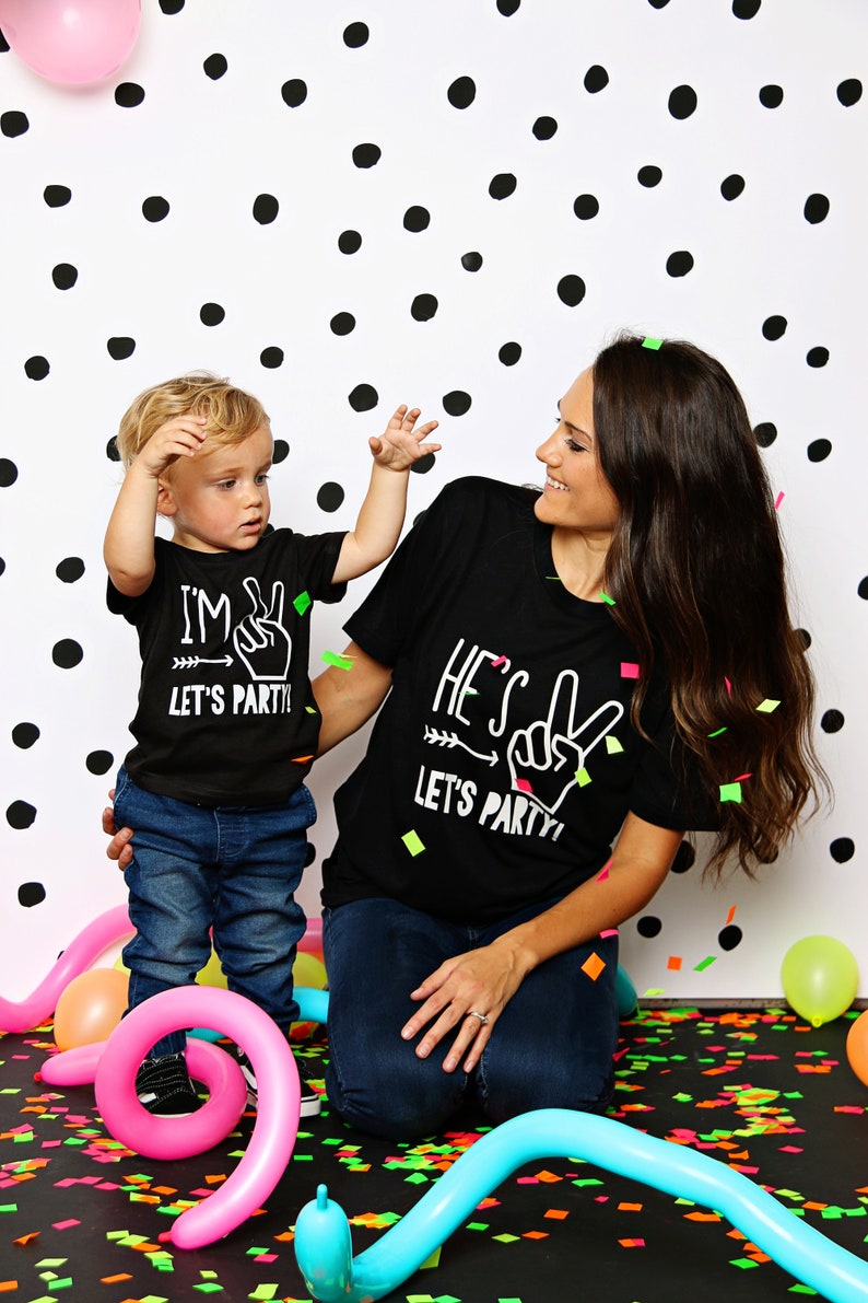 May include: A mother and child wearing matching black t-shirts with white text that says "I'm 2 Let's Party!" and "He's 2 Let's Party!" The child is holding up two fingers in a peace sign. They are standing in front of a black and white polka dot background with colorful confetti on the floor.