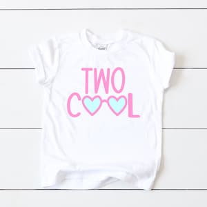 May include: White t-shirt with pink text that says "TWO COOL" with blue heart-shaped sunglasses.