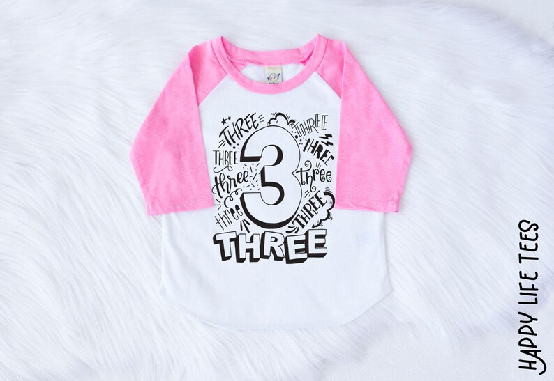 May include: A white and pink baseball tee shirt with a black graphic that says "Three" with the number 3 in the center. The number 3 is surrounded by the word "Three" written multiple times.